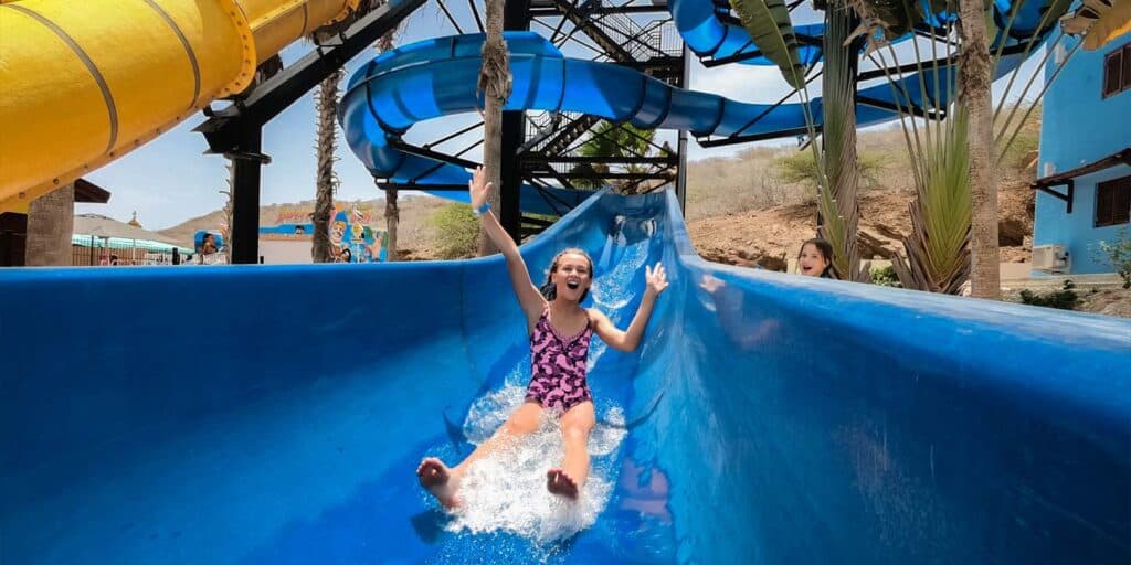 Een lachend kind in een paars badpak glijdt van een felblauwe waterglijbaan bij Dagpas Kunuku Aqua Resort, armen omhoog en spetterend onder de zonnige hemel terwijl een ander kind met een Dagpas toekijkt vanaf de rand bij palmbomen en nog meer glijbanen.