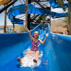 At Daypass Kunuku Aqua Resort, a girl in a purple patterned swimsuit smiles with arms raised as she slides down a bright blue water slide. Water splashes at her feet, palm trees and twisting slides surround the sunny scene. Another child is partly visible behind her.