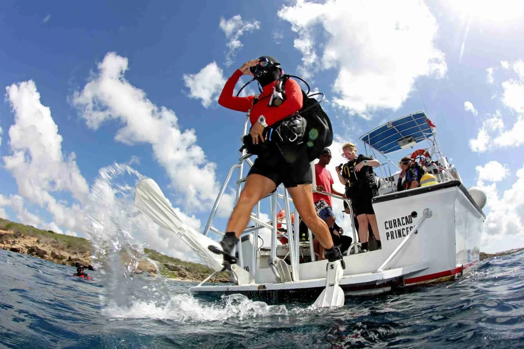 A diver in full gear steps off the boat for a TWO TANK AM BOAT DIVING trip as others watch from the sunlit, partly cloudy deck with the shoreline visible in the distance.
