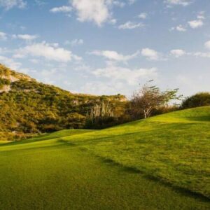 A golfer in pink and white swings on a lush green course, sand spraying under a blue sky with clouds—ideal for the 1 Day Golf Tour and Clinic amid rolling hills and rocky outcrops.