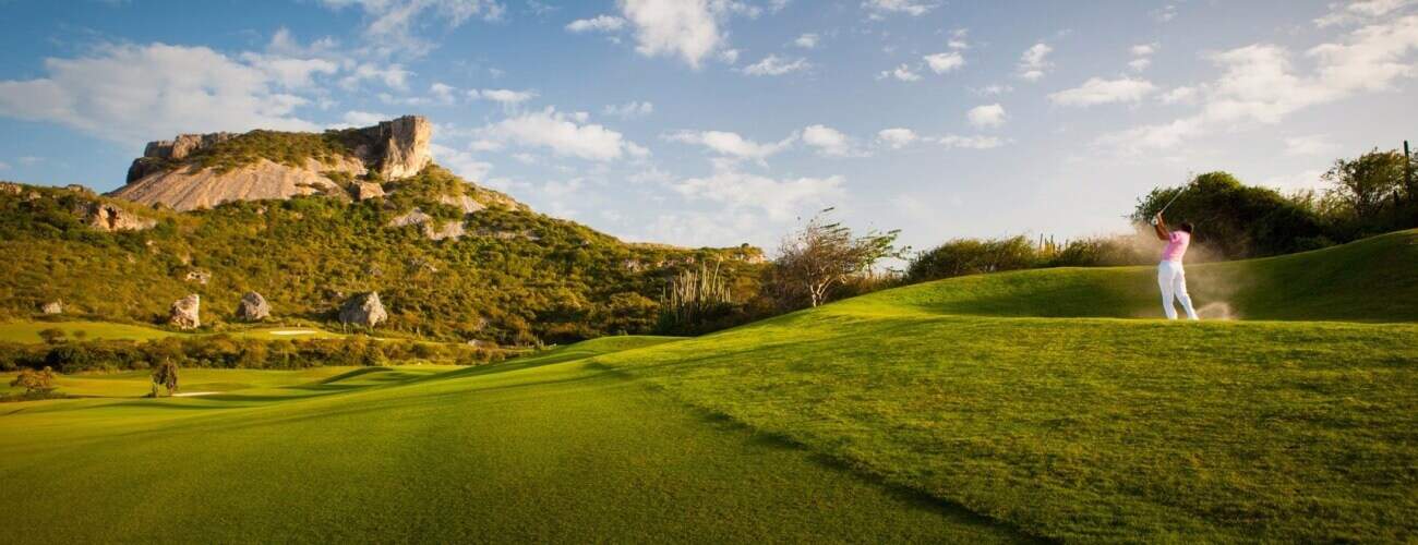 A golfer in pink and white swings on a lush green course, sand spraying under a blue sky with clouds—ideal for the 1 Day Golf Tour and Clinic amid rolling hills and rocky outcrops.