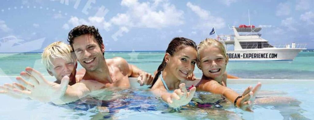 A smiling family of four—two adults and two kids—swims in clear blue water, reaching toward the camera on their 100% Island Adventure trip. Behind them, a white boat with CARIBBEAN-EXPERIENCE.COM floats on the turquoise sea.
