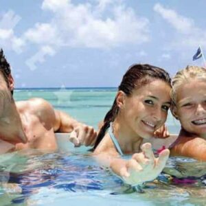 A smiling family of four—two adults and two kids—swims in clear blue water, reaching toward the camera on their 100% Island Adventure trip. Behind them, a white boat with CARIBBEAN-EXPERIENCE.COM floats on the turquoise sea.