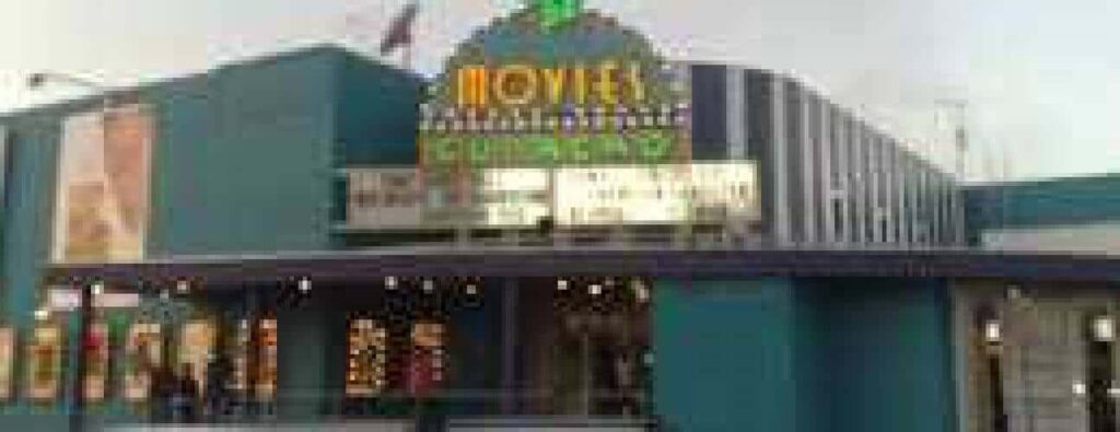 A festive teal theater decorated with lights features a brightly lit MOVIES marquee and multiple front windows. Poster displays are on the left, promoting the 2 for 1 movies special.