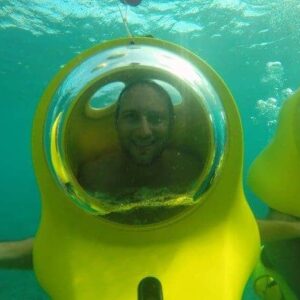 A man smiles inside a yellow Aquafari Excursion underwater scooter with a clear bubble helmet, submerged in turquoise water. Another scooter appears to the right, while a hand is extended from the left side of the image.