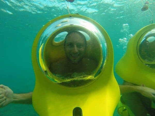 A man smiles inside a yellow Aquafari Excursion underwater scooter with a clear bubble helmet, submerged in turquoise water. Another scooter appears to the right, while a hand is extended from the left side of the image.