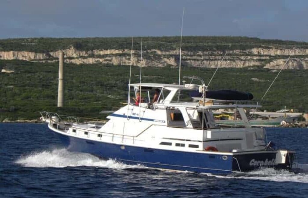 A white and blue motor yacht cruises on deep blue water near cliffs and greenery. Enjoy a Deep Sea Fishing Charter (Half Day) as industrial buildings and a tall smokestack rise in the background.