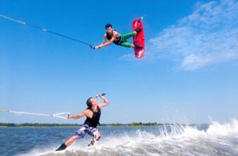 Two people are wakeboarding on a lake. One launches into an aerial trick, holding a red board and rope high above the water, while the other rides the Curacao-blue wake below, sipping from a bottle with trees and sky in the background.