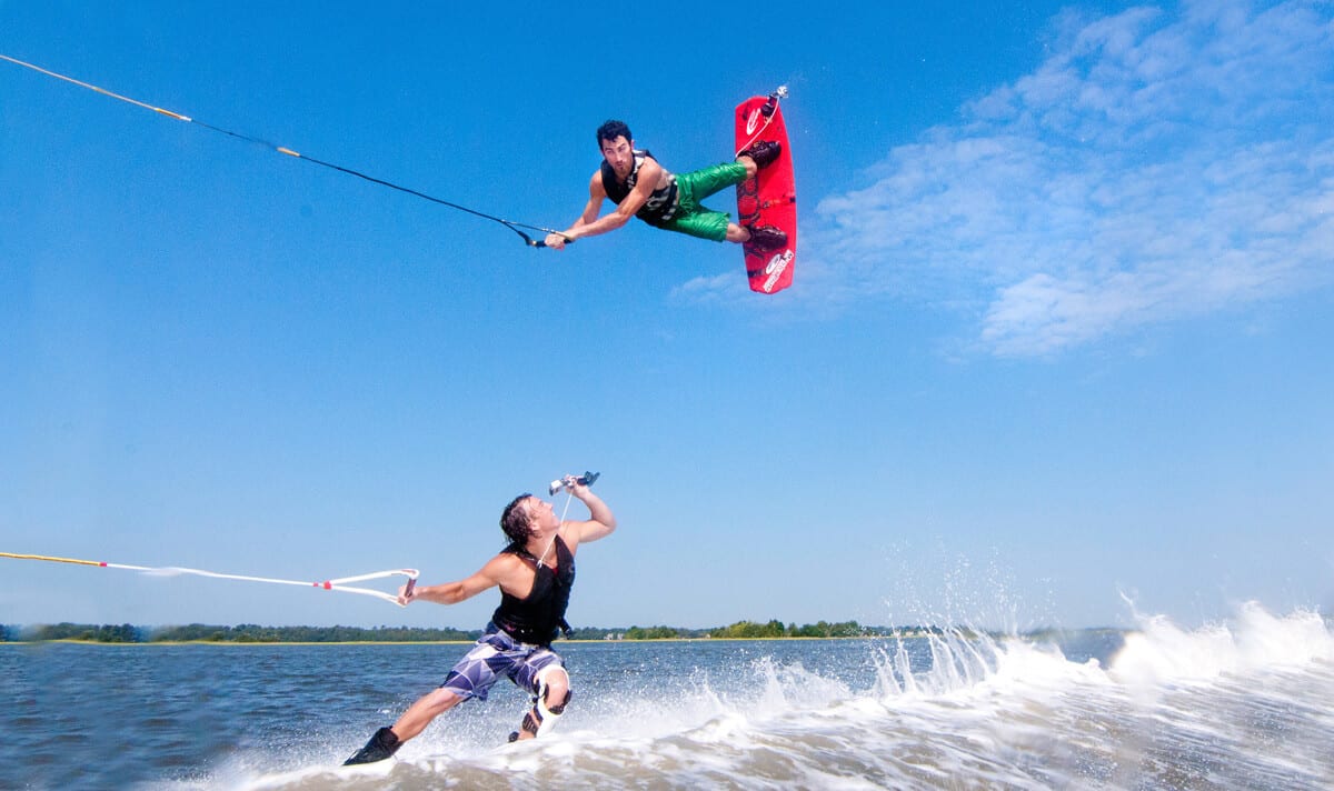 Two people are wakeboarding on a lake. One launches into an aerial trick, holding a red board and rope high above the water, while the other rides the Curacao-blue wake below, sipping from a bottle with trees and sky in the background.