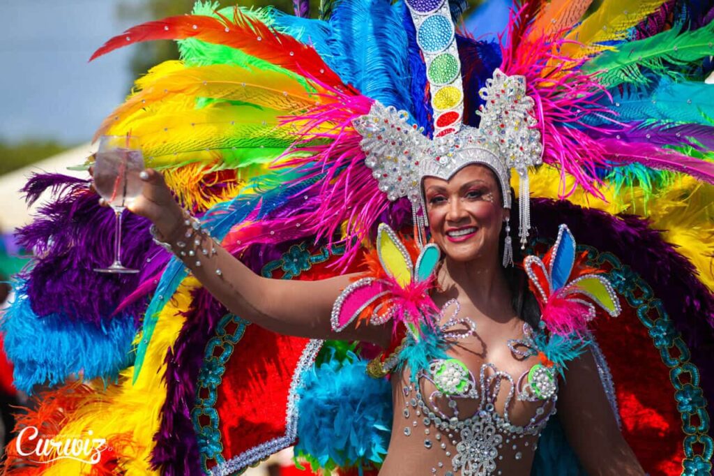 A woman in a vibrant, elaborate Curacao carnival costume with sparkling jewels, colorful feathers, and butterfly decorations smiles and holds up a glass. She wears a detailed headdress and festive makeup, celebrating outdoors during a lively parade.