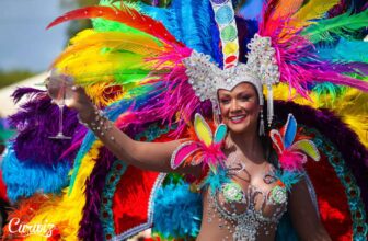A woman in a vibrant, elaborate Curacao carnival costume with sparkling jewels, colorful feathers, and butterfly decorations smiles and holds up a glass. She wears a detailed headdress and festive makeup, celebrating outdoors during a lively parade.