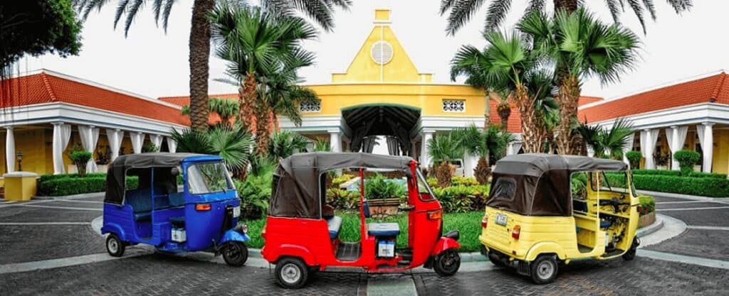 Three vibrant tuk-tuks—blue, red, and yellow—are parked together on a palm-lined driveway in front of a yellow building with white columns and a red roof, ready to start the TukTuk City Tour.