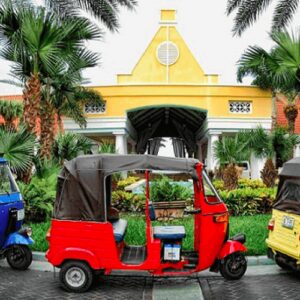 Three vibrant tuk-tuks—blue, red, and yellow—are parked together on a palm-lined driveway in front of a yellow building with white columns and a red roof, ready to start the TukTuk City Tour.