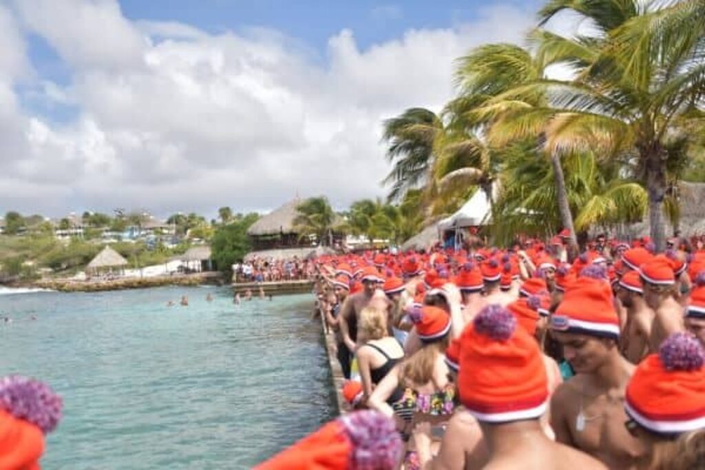 A large group of people in swimsuits and red winter hats with pom-poms stand by the edge of a tropical beach in Curacao, palm trees behind them, preparing for a swim. The sky is partly cloudy, the water is clear blue, and more people watch from the shore.