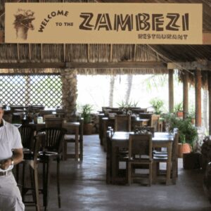 A man in a white shirt and cap smiles at the entrance of Zambezi Restaurant, an open-air spot with a thatched roof—ideal for relaxing after your African Night Special Tour at Ostrich farm.