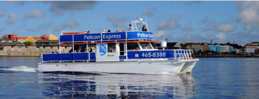 De witte en blauwe Pelican Express veerboot vervoert passagiers voor de Snorkeltour met gidsen. Op de achtergrond zie je kleurrijke gebouwen, een stenen muur en een gedeeltelijk bewolkte lucht. Op de veerboot staat dit telefoonnummer: 465-8388.