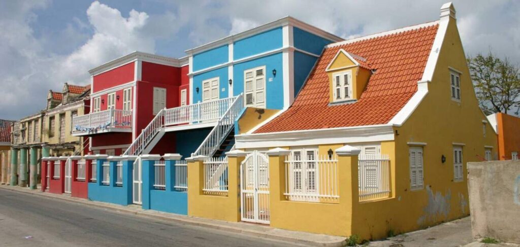 A row of colorful houses reminiscent of Curacao lines a quiet street. Painted in bright red, blue, and yellow with white shutters and orange tiled roofs, the scene is lively beneath a partly cloudy sky.