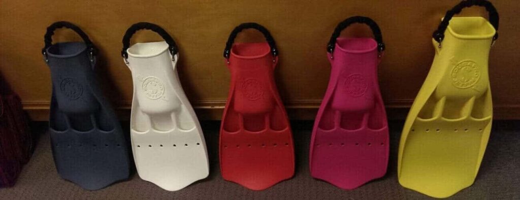 Five Guided Shore Dive fins in black, white, red, pink, and yellow stand upright side by side on a carpeted floor against a brown wall. Each features a wide foot pocket and a thick black strap at the heel.