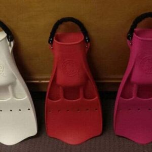 Five Guided Shore Dive fins in black, white, red, pink, and yellow stand upright side by side on a carpeted floor against a brown wall. Each features a wide foot pocket and a thick black strap at the heel.