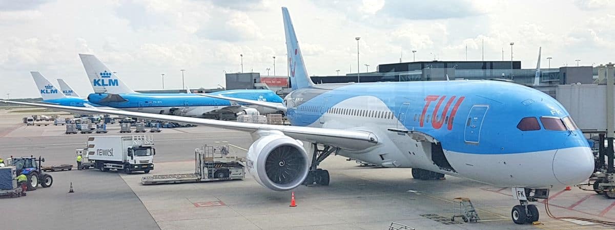 A TUI airplane is parked at an airport gate in Curacao with its engine and nose visible. Two KLM planes are seen in the background, while ground crew, luggage carts, and airport equipment are present on the tarmac under a cloudy sky.
