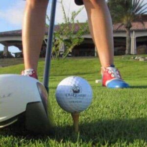 A close-up of a golf club and ball on a tee, ready for action with the Advanced 2 day Package. The golfer’s colorful shoes and legs are visible on the green course, framed by palm trees and a tiled-roof building under sunny skies.