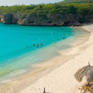A few people enjoy All West Beach Hopping on a tropical beach with white sand, turquoise water, palm trees, two thatched-roof huts, and lush green cliffs under a sunny sky.