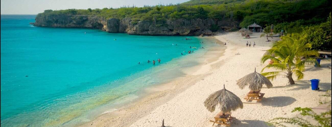 Een tropisch strand met wit zand en turquoise water, waar mensen zwemmen in de buurt van palapa parasols en picknicktafels - All West strandhoppen is ideaal om te ontspannen, omringd door weelderige bomen en kliffen onder een heldere hemel.