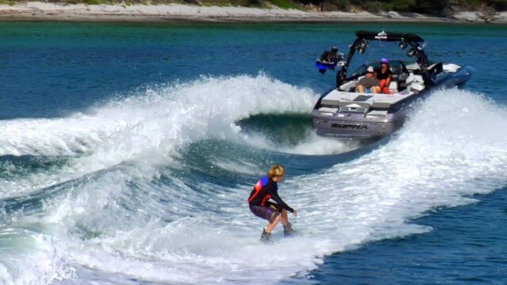 A person in a wetsuit wakeboards on a large wave created by the All-inclusive watersport with three people onboard, gliding through clear blue water near a tree- and sand-lined shore.