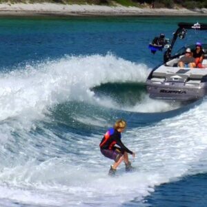 A person in a wetsuit wakeboards on a large wave created by the All-inclusive watersport with three people onboard, gliding through clear blue water near a tree- and sand-lined shore.