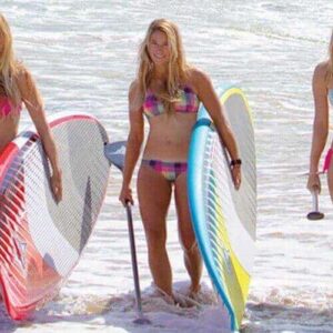 Three women enjoy Beach Hopping with SUP, standing in shallow ocean water with paddleboards and smiling as they come ashore, waves lapping their feet and sandy beach ahead.