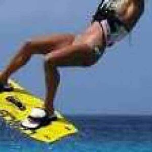During Beginner Lesson 1 - Kite Flying Basics, a person in a harness and swimsuit soars above the ocean, gripping a yellow wakeboard with both feet strapped in—capturing an exciting moment under clear blue skies.