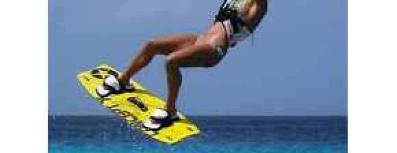 During Beginner Lesson 1 - Kite Flying Basics, a person in a harness and swimsuit soars above the ocean, gripping a yellow wakeboard with both feet strapped in—capturing an exciting moment under clear blue skies.