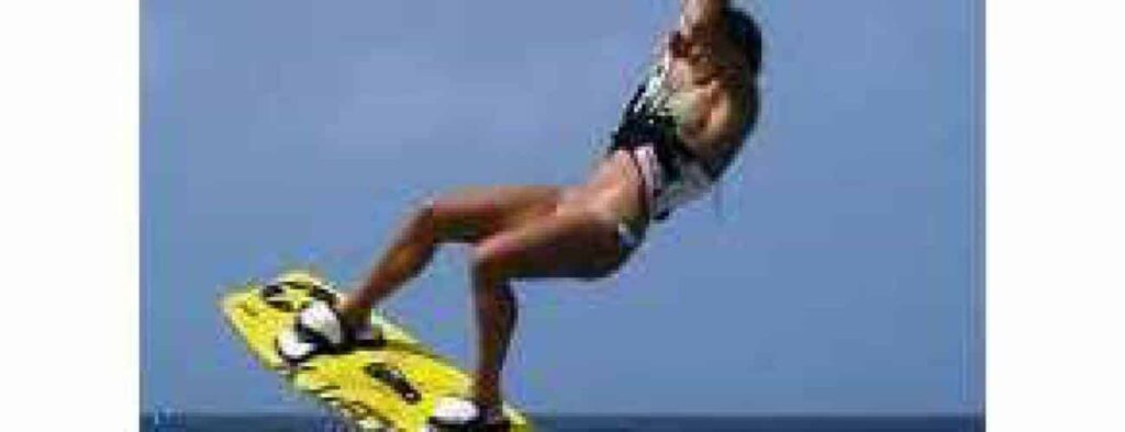 A person wakeboards mid-air above water under a clear blue sky, holding a rope and showcasing skills learned in "Beginner lesson 2 - Core Skills," using a bright yellow board while wearing a swimsuit and life vest.