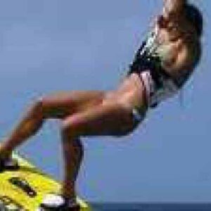 A person wakeboards mid-air above water under a clear blue sky, holding a rope and showcasing skills learned in "Beginner lesson 2 - Core Skills," using a bright yellow board while wearing a swimsuit and life vest.