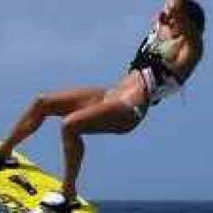 Una persona en bañador hace kitesurf sobre el océano, demostrando "Lección para principiantes 3 - Comienzo en el agua" con una tabla amarilla con arnés, elevándose bajo el cielo azul en esta dinámica escena de deportes acuáticos.