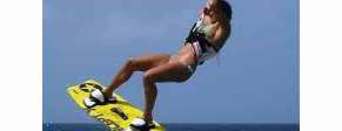 A person is mid-air kiteboarding over the ocean during Beginner Lesson 3 - Water Start, holding the harness with a bright yellow board and a clear blue sky in the background.