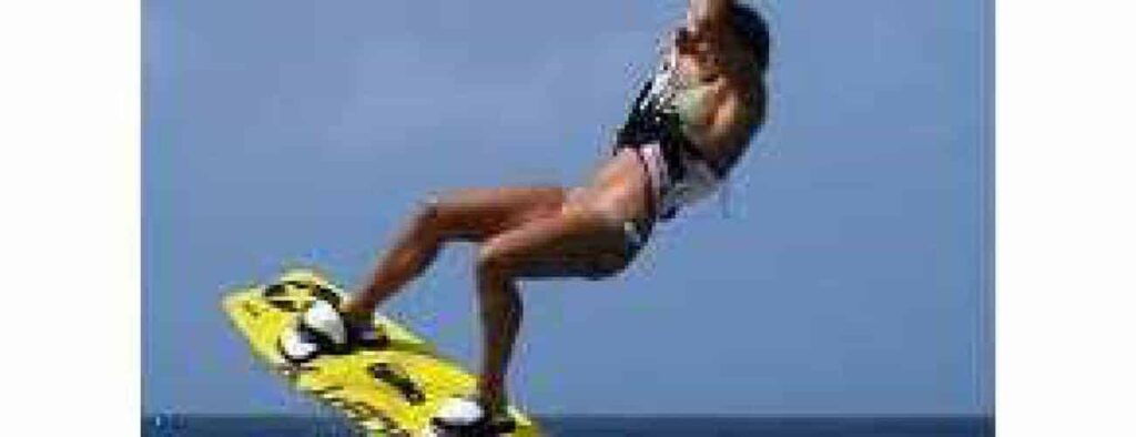 In Beginner lesson 4 – Water Start & Riding, a wakeboarder in a swimsuit and life vest soars mid-air on a yellow board against a clear blue sky, capturing the energy and excitement of the moment.