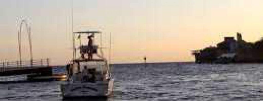 A small white boat with several people aboard, part of Big Game Fishing Trips, floats on calm water at sunset. The sky glows orange, rocky shoreline is silhouetted right, and a dock stands to the left.