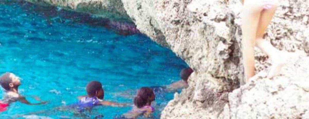 Four people swim in clear, blue water near jagged rocks, channeling adventure and natural beauty in the Blue room cycle adventure. On the right, a person in a pink swimsuit climbs the rocks, adding excitement to this vibrant outdoor scene.
