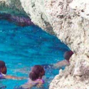 Four people swim in clear, blue water near jagged rocks, channeling adventure and natural beauty in the Blue room cycle adventure. On the right, a person in a pink swimsuit climbs the rocks, adding excitement to this vibrant outdoor scene.