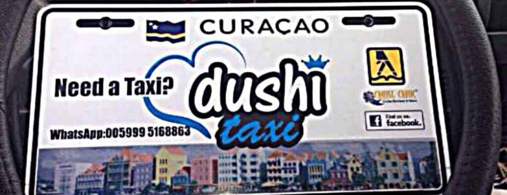 Een Curaçaos taxibord voor Taxi naar Boca Tabla toont "Taxi nodig? Dushi taxi," WhatsApp contact, Facebook logo, kleurrijke gebouwen eronder, en bovenop de Curaçaose vlag, "CURACAO," en logo's van taxiservices voor naadloos reizen.