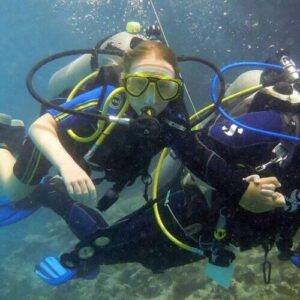 Two scuba divers in wetsuits, masks, fins, and oxygen tanks swim hand-in-hand above a coral reef. Both wear yellow-blue gear while enjoying the Bubblemaker diving Program amid bubbles and clear blue water.