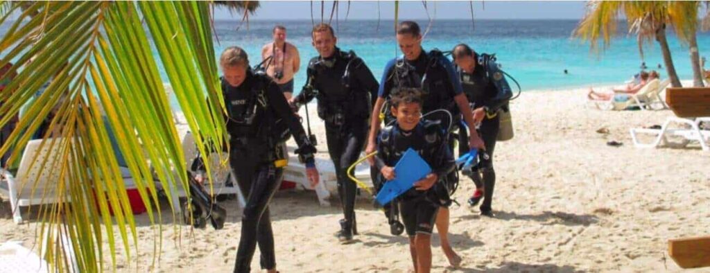 Five kids (ages 8-10) in wetsuits and scuba gear, part of the PADI Bubblemaker program, walk from a sunny tropical beach toward the camera, smiling. Palm fronds, lounge chairs, and turquoise ocean complete the vibrant scene.