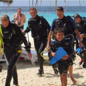 Five kids (ages 8-10) in wetsuits and scuba gear, part of the PADI Bubblemaker program, walk from a sunny tropical beach toward the camera, smiling. Palm fronds, lounge chairs, and turquoise ocean complete the vibrant scene.