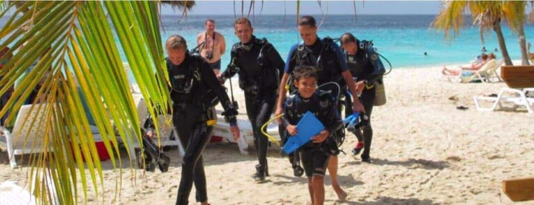Five kids (ages 8-10) in wetsuits and scuba gear, part of the PADI Bubblemaker program, walk from a sunny tropical beach toward the camera, smiling. Palm fronds, lounge chairs, and turquoise ocean complete the vibrant scene.