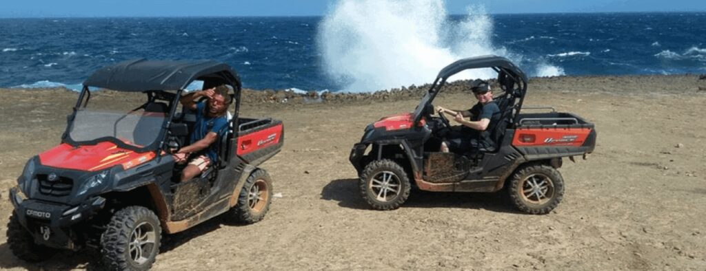 Two people enjoy a Buggy Tour in separate red off-road vehicles by a rocky coastline. Ocean waves crash against the rocks, sending white spray into the air beneath a clear sky, while the ground remains dry and rugged.