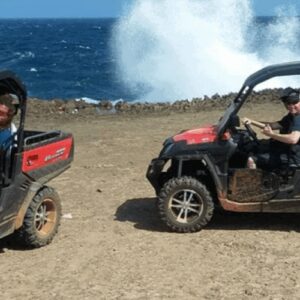 Two people enjoy a Buggy Tour in separate red off-road vehicles by a rocky coastline. Ocean waves crash against the rocks, sending white spray into the air beneath a clear sky, while the ground remains dry and rugged.