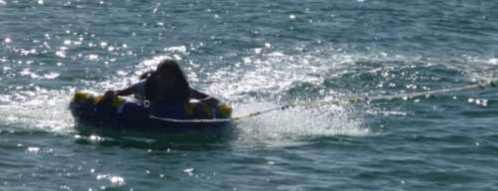 Iemand rijdt op een opblaasbare buis van Botsband die aan een touw wordt voortgetrokken over zonovergoten, kabbelend water. Rechtop zittend op de buis creëren ze een kielzog achter zich terwijl het zonlicht schittert op het wateroppervlak.