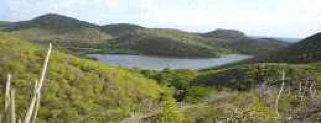 A Camping scene unfolds with green, rolling hills centered around a small lake. The lush spring or summer vegetation and clear sky are complemented by background hills, partially covered in shrubs and trees.