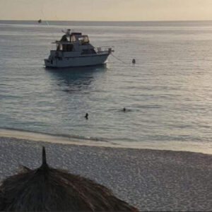 Kamperen op Klein Curaçao biedt een serene zonsondergang op het strand met een kalme zee, kleine bootjes, een jacht voor de kust en mensen in silhouetten aan de rand van het water. Gezellige palapahutten, ideaal voor kamperen, staan verspreid over de zandkust.